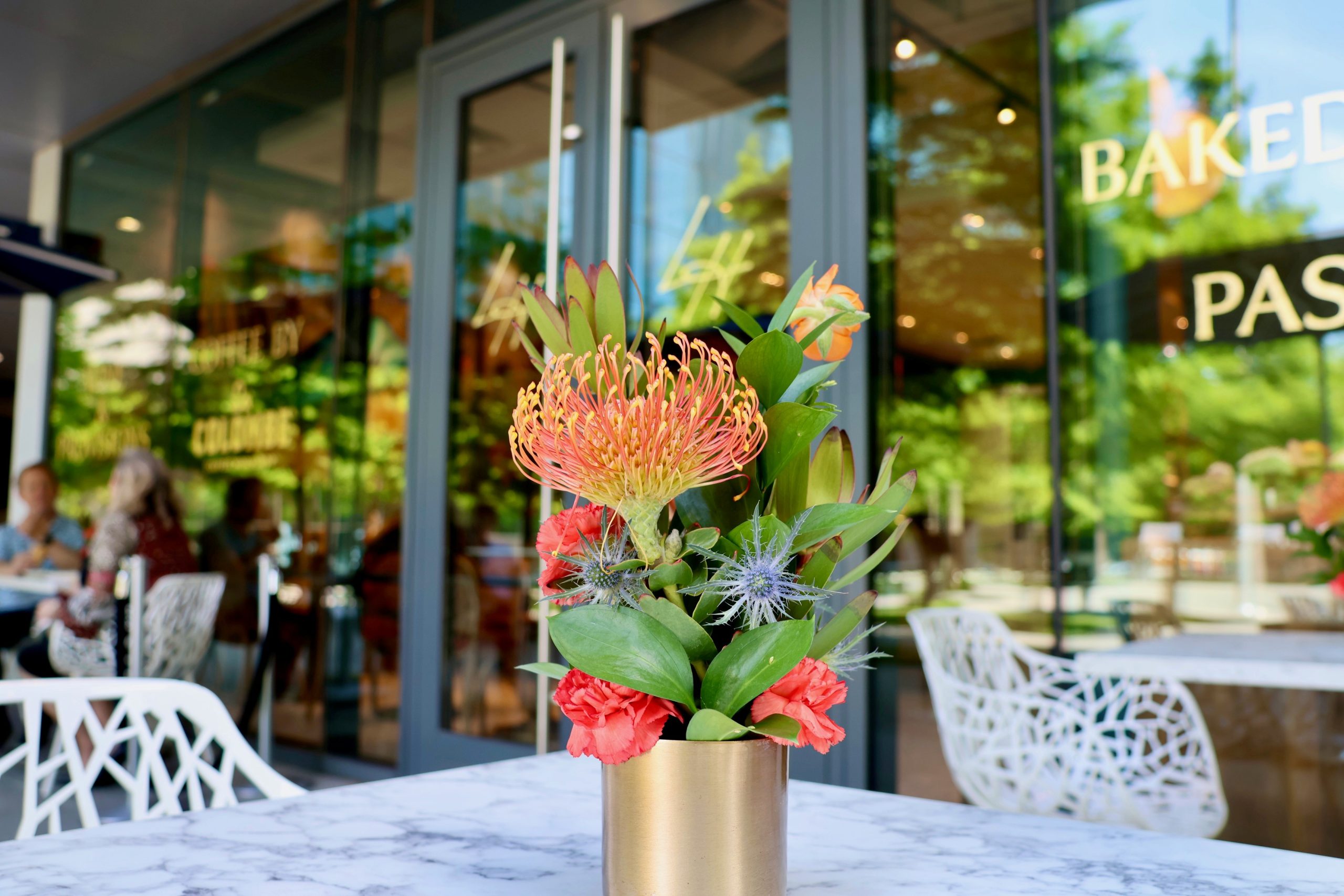 Little Heron - A small floral arrangement in a gold vase sits on a marble table outside a café, with people dining in the background and large glass windows reflecting greenery.